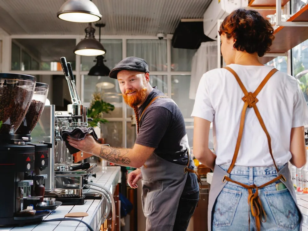 A barista wiping down the espresso machine behind the counter of a small coffee shop